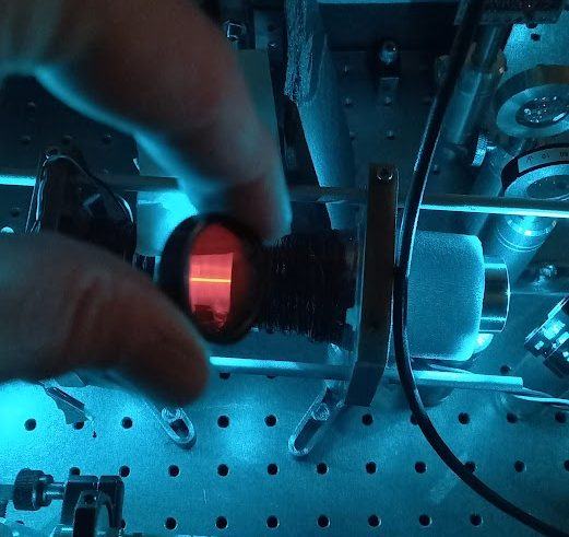 A hand holds a small glass vapour cell glowing red, in front of optics lit in blue